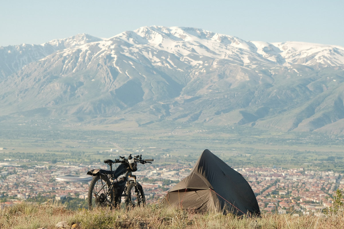 Elliot Silander's tent and loaded touring bicycle pitched on a ridge above the Erzincan valley in Turkey, with snow-capped mountains behind.