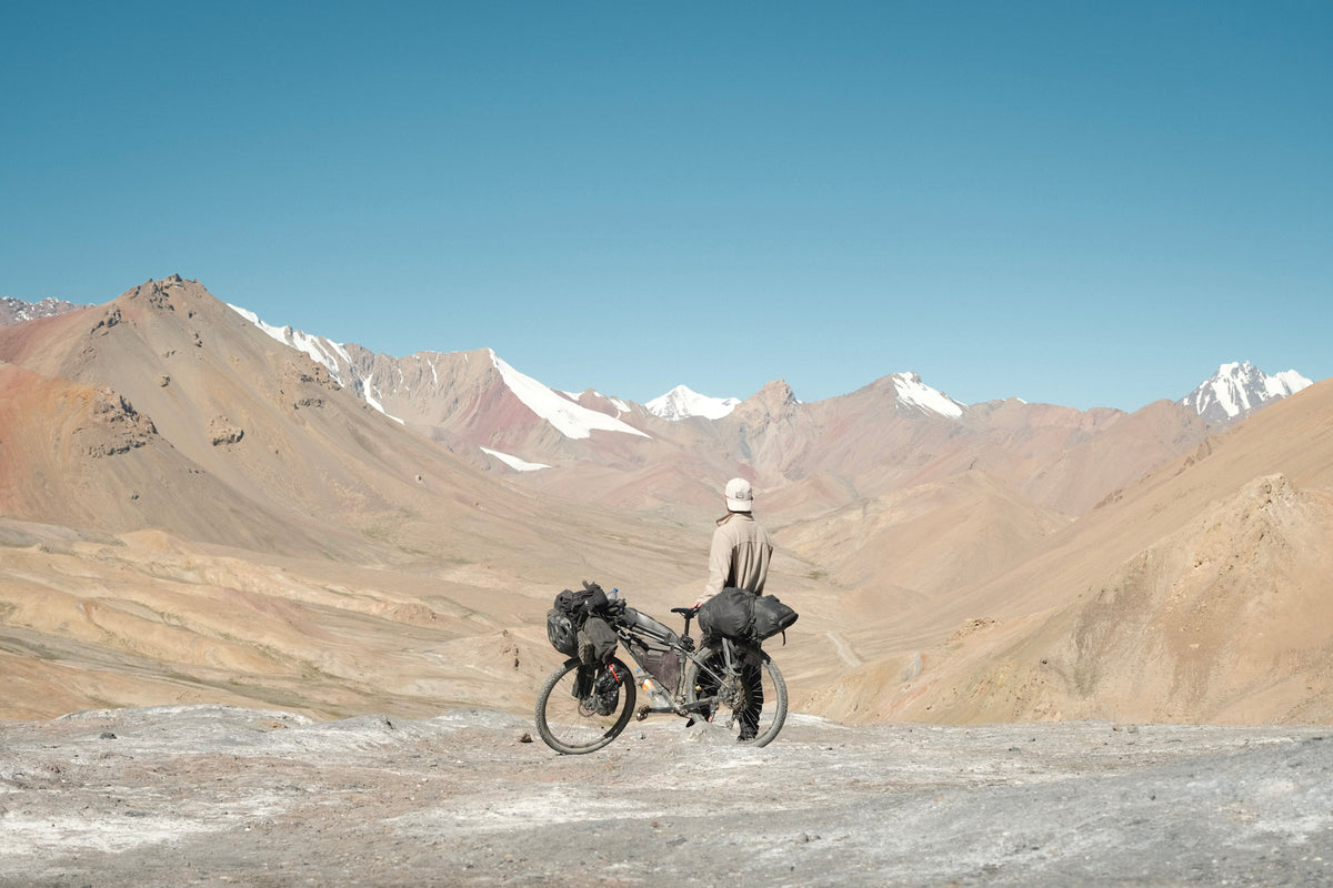 Elliot Silander with his loaded touring bicycle on top of the Ak-Baital pass in Tajikistan, snow-capped mountains behind.